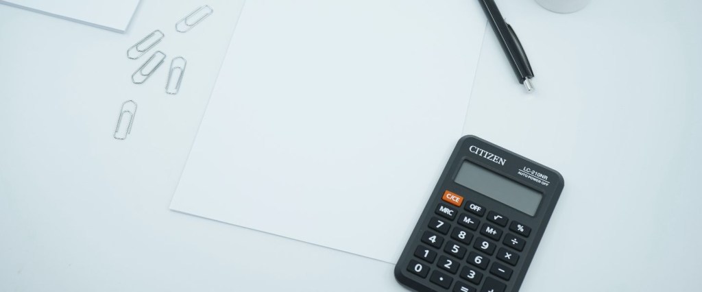 Endividamento e inadimplência: imagem de uma calculadora preta sobre uma mesa branca com folha de papel, clips, caneta e vaso de suculenta simbolizando contabilidade de dívidas. Foto: @mediamodifier/Unsplash