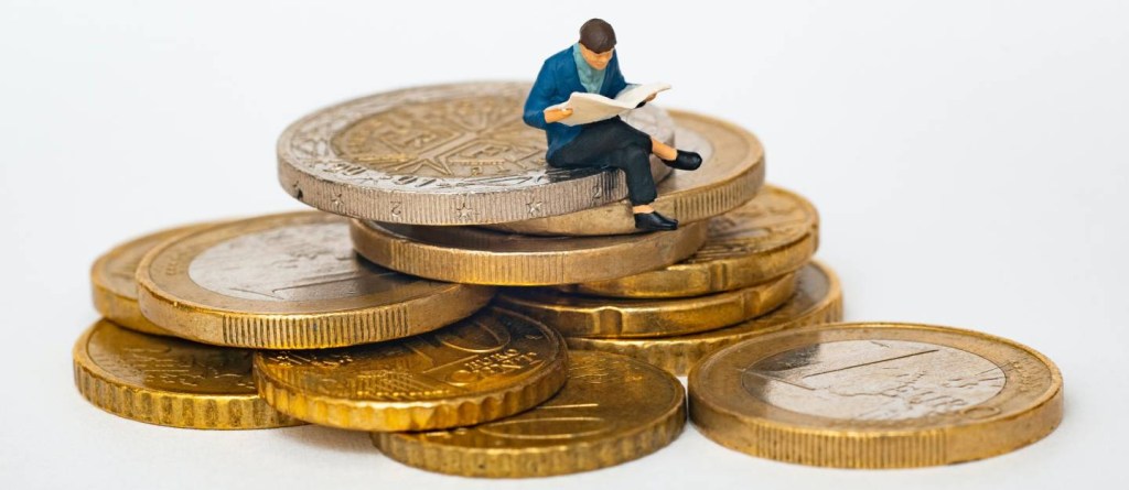 Imagem mostra pequeno boneco de um homem sentado sobre uma pilha de moedas enquanto lê um jornal - Como declarar contas bancárias