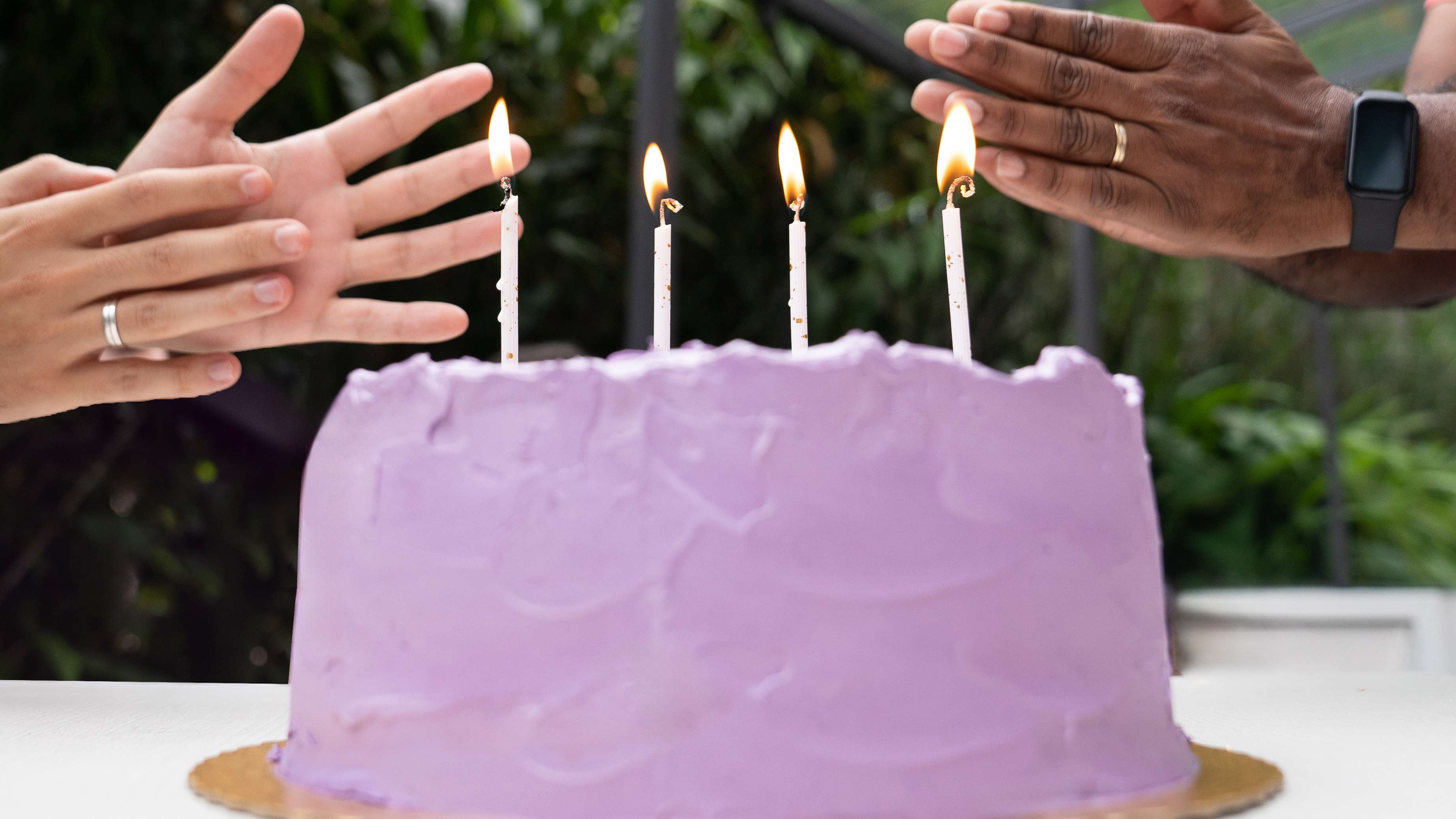 Na imagem aparece um bolo com cobertura roxa e algumas velas com duas mãos, à esquerda e à direita, batendo palmas