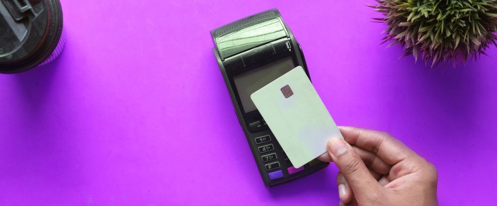 Mão masculina aproxima um cartão branco da maquininha por contactless. o fundo é roxo. foto: @Towfiqubarbhuiya/Unsplash