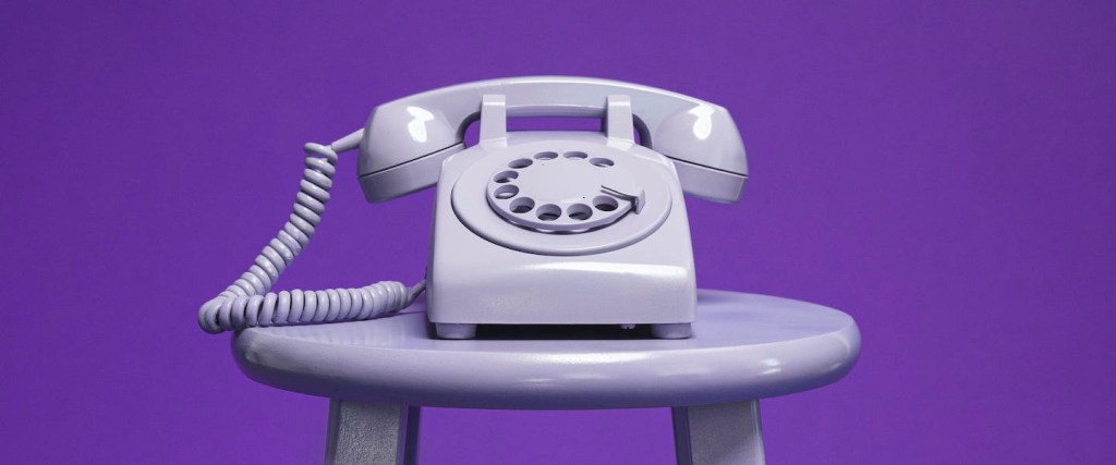 Imagem de um telefone antigo e roxo posicionado sobre um banquinho de madeira da mesma cor.
