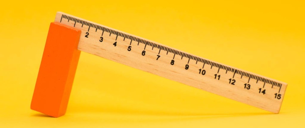 Imagem mostra uma régua de madeira de 15 centímetros com um cabo vermelho. Ela está sobre um fundo amarelo - valuation