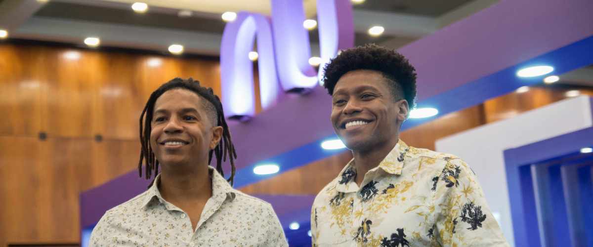 Na imagem, aparecem os dois fundadores da AfroSaúde. À esquerda Igor, um homem negro de cabelos trançados, aparece sorrindo. Ao lado dele, à direita, Arthur, que tem o cabelo afro e também sorri. Ao fundo, o logo do Nubank