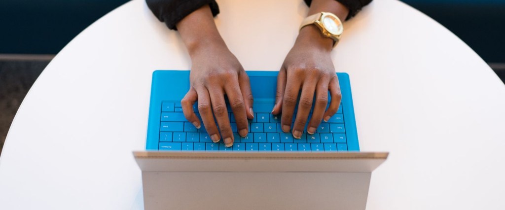 Empreendedorismo negro: mulher negra tecla em notebook azul com um relógio de pulso no braço esquerdo. O notebook está sobre uma mesa branca. Foto: @WOCInTech/Nappy