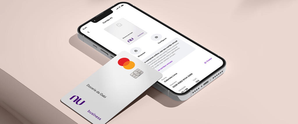 Cartão de Crédito PJ: imagem do novo modelo de cartão da conta PJ. Ele é prateado, com o logo do Nubank em roxo. Também é possível observar um celular ligado com a tela do app do Nubank.