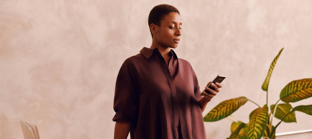 Imagem mostra uma mulher negra de cabelos curtos usando um celular. Ela veste uma blusa marrom.