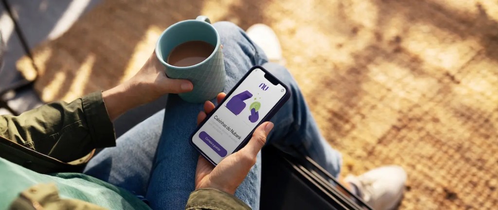 Foto destaca a mão de uma pessoa segurando um celular, com a tela para o app do Nubank, e com a outra mão segurando uma caneca com café.