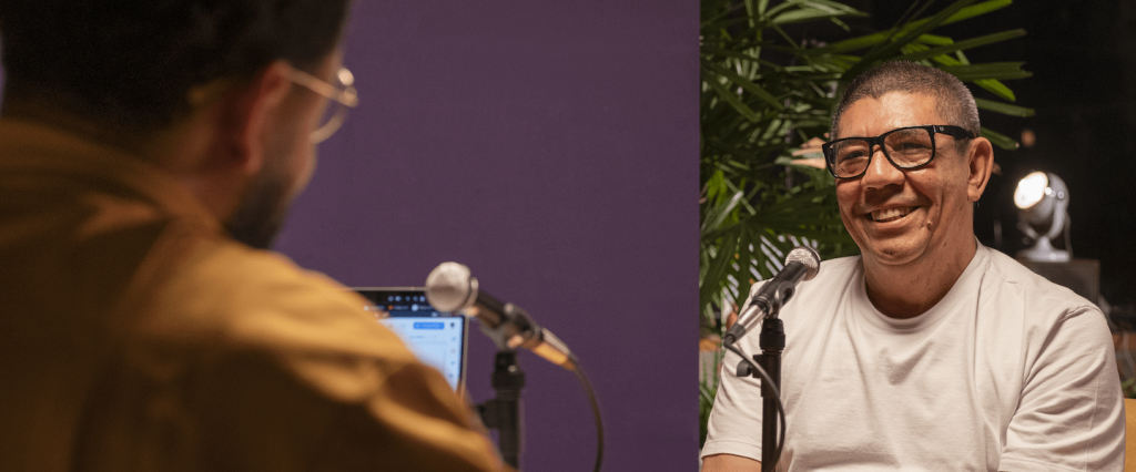 Emicida e Sergio Vaz de frente um para o outro, na mesa do podcast Chamaê. No ângulo da imagem, Emicida está de costas, usando uma jaqueta bege. Sergio está sorrindo, usa óculos e uma camiseta branca.