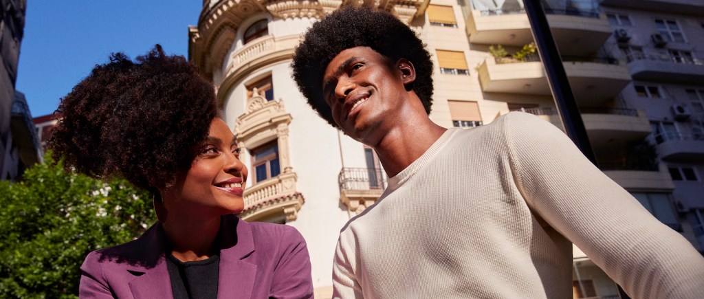 Foto mostra um casal formado por uma mulher preta e um homem preto, ambos de cabelo crespo black. Ela está com um paletó roxo e ele veste uma blusa de malha bege. Ambos estão numa viagem.