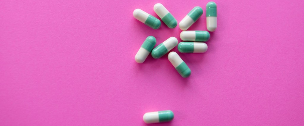Um punhado de cápsulas de remédios nas cores branco e verde estão dispostos sobre um fundo pink.