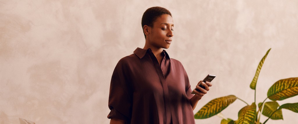 Imagem mostra uma mulher negra de cabelos curtos usando um celular. Ela veste uma blusa marrom.