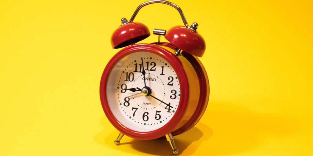 Foto de um pequeno relógio despertador vermelho sobre um fundo amarelo.