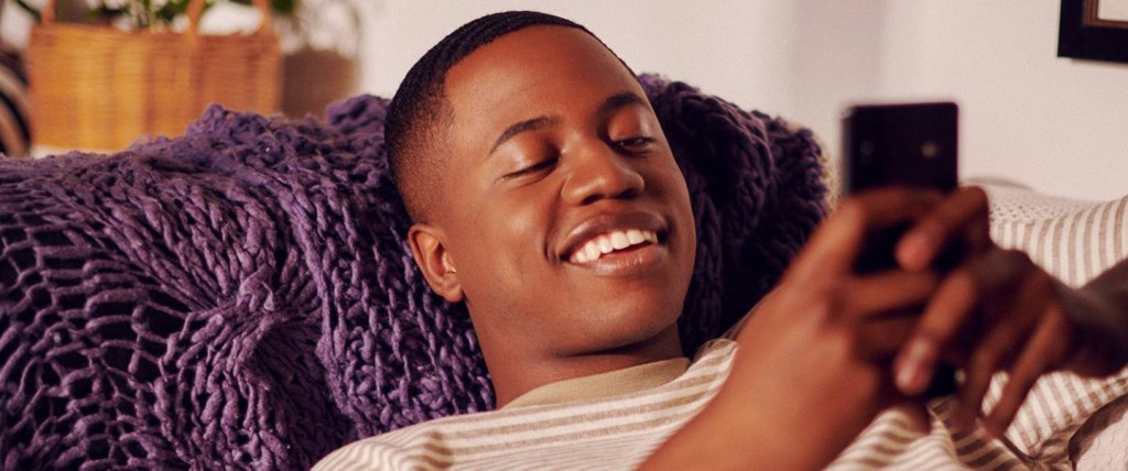 Jovem negro de cabelos curtos está deitado e sorrindo enquanto usa o celular.