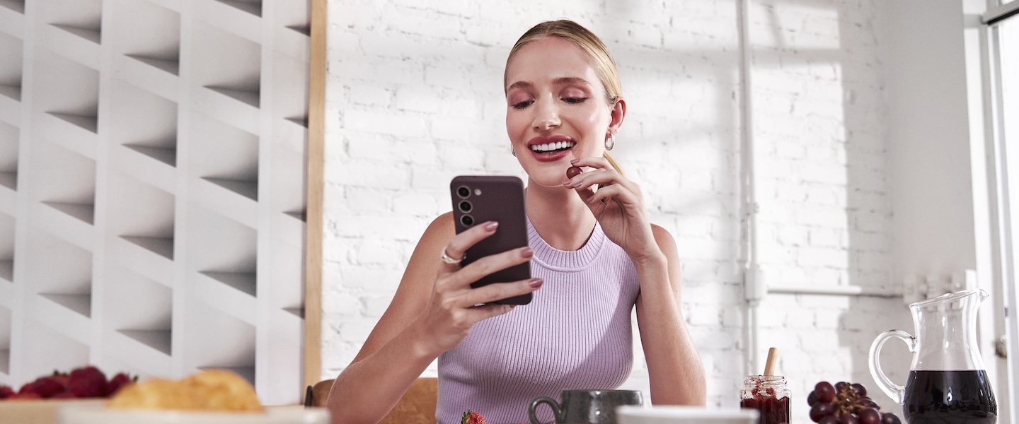 A imagem mostra uma pessoa vestindo uma blusa lilás, sentada à mesa de um café, aparentemente distraída com um celular em suas mãos. O ambiente em volta parece moderno, com paredes brancas de tijolos à vista e uma janela grande fornecendo luz natural. Sobre a mesa há alguns itens de café da manhã ou lanche, como uma jarra com bebida escura (possivelmente café ou suco), um pote de geleia, uvas e um prato com o que parece ser um croissant e frutas.