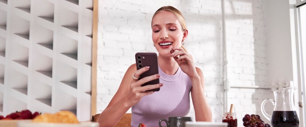 A imagem mostra uma pessoa vestindo uma blusa lilás, sentada à mesa de um café, aparentemente distraída com um celular em suas mãos. O ambiente em volta parece moderno, com paredes brancas de tijolos à vista e uma janela grande fornecendo luz natural. Sobre a mesa há alguns itens de café da manhã ou lanche, como uma jarra com bebida escura (possivelmente café ou suco), um pote de geleia, uvas e um prato com o que parece ser um croissant e frutas.