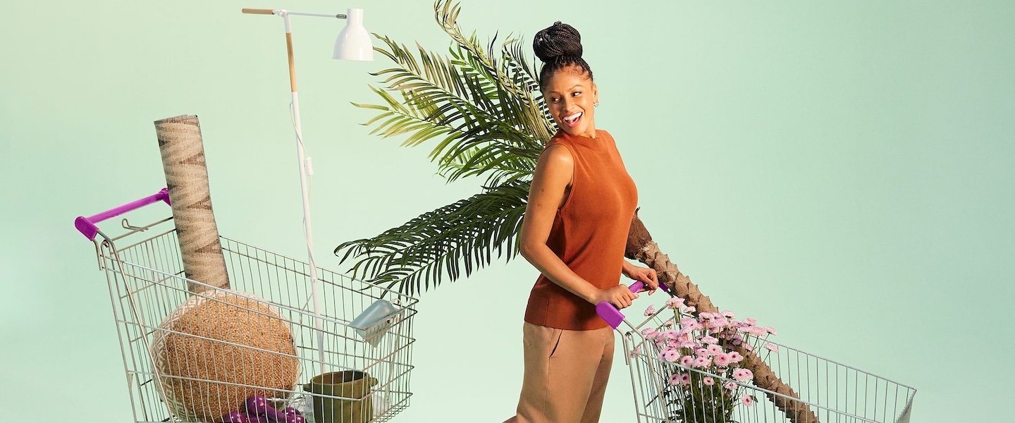 imagem de uma mulher negra empurrando um carrinho de compras num cenário de fundo verde. Ao lado dela, há um outro carrinho. Ambos estão com objetos dentro dele. A mulher usa uma roupa marrom e o cabelo está preso em um coque bem alto.