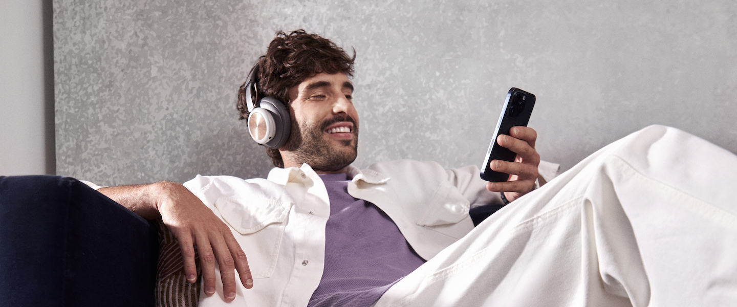 Um homem branco sentado confortavelmente em um sofá, sorrindo enquanto usa um headset e olha para o celular em uma de suas mãos.