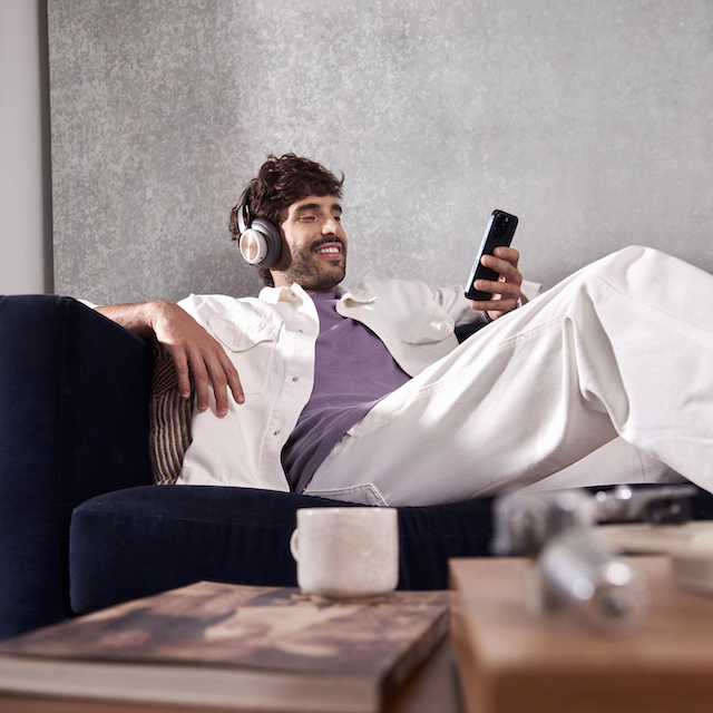 Um homem branco sentado confortavelmente em um sofá, sorrindo enquanto usa um headset e olha para o celular em uma de suas mãos.