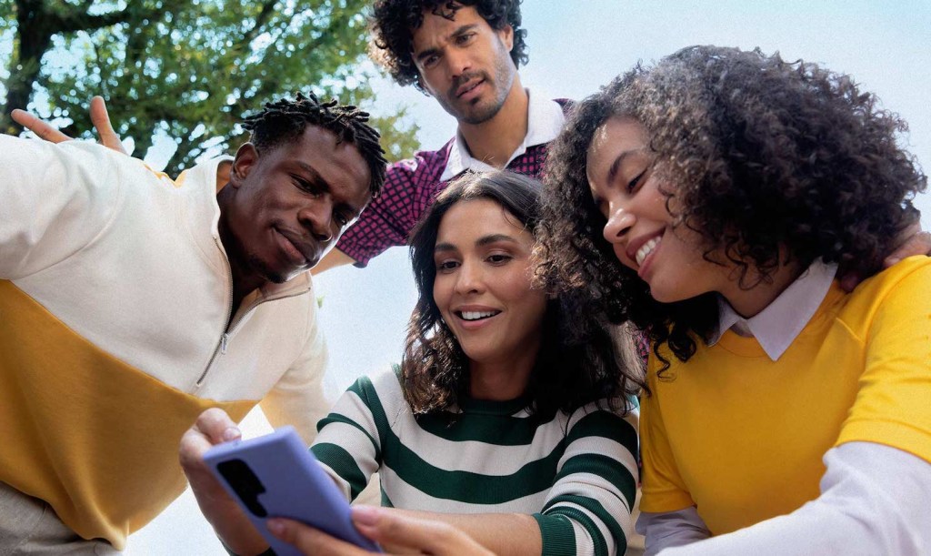 foto de um grupo de amigos (duas mulheres e. dois homens),uma está com o celular na mão mostrando algo para eles.