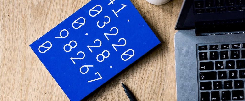 Imagem de um caderninho azul, com números na capa. Ao lado dele há um notebook e uma caneta. Os objetos estão sobre uma mesa de madeira.