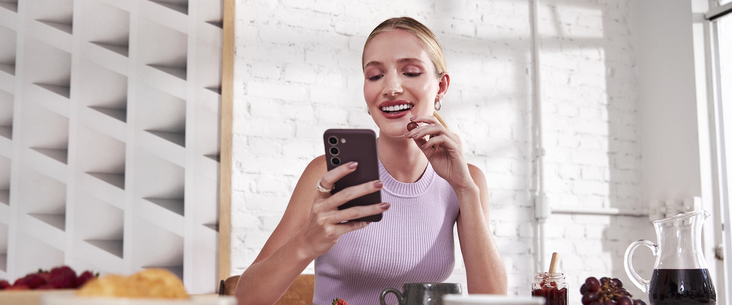 Imagem de uma mulher loira sorrindo, comendo um morango. Ela está de cabelo preso, usando uma blusa regata lilás e segurando um celular em uma das mãos. Ela está à mesa, com várias bebidas e comidas.