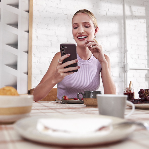 Imagem de uma mulher loira sorrindo, comendo um morango. Ela está de cabelo preso, usando uma blusa regata lilás e segurando um celular em uma das mãos. Ela está à mesa, com várias bebidas e comidas.