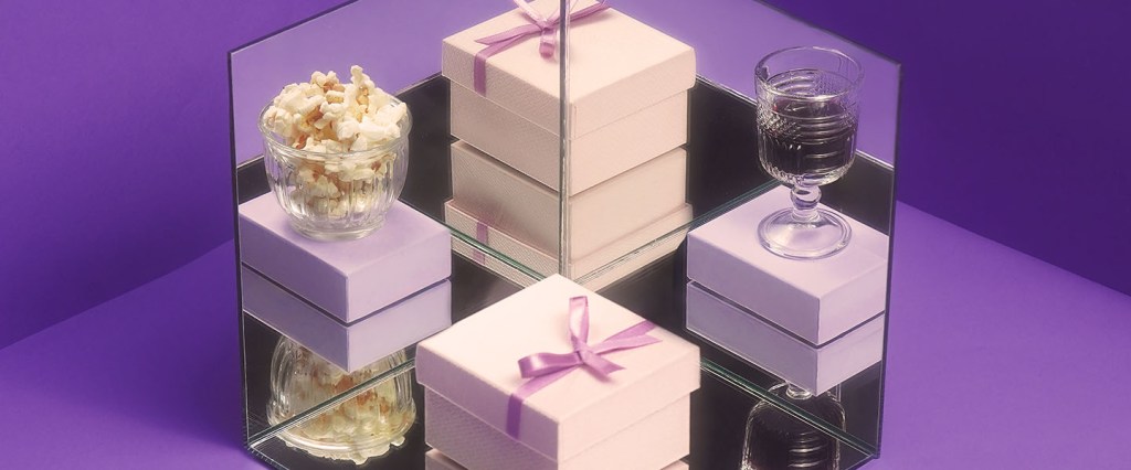 Presente de Natal: imagem de caixas de presente nas cores rosa e roxo. Sobre uma delas há uma taça com bebida e, em outra, um potinho de pipoca.