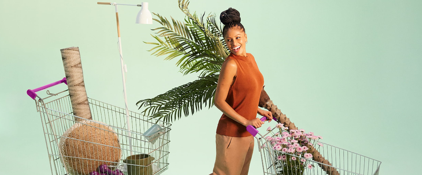 Imagem de uma mulher negra empurrando um carrinho de compras num cenário de fundo verde. Ao lado dela, há um outro carrinho. Ambos estão com objetos dentro dele. A mulher usa uma roupa marrom e o cabelo está preso em um coque bem alto.