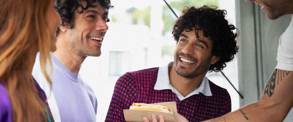 Imagem mostra três pessoas em frente conversando: uma mulher branca, de cabelos longos avermelhados e casaco roxo escuro, um homem branco com camiseta roxa clara e outro homem branco, com barba e camiseta roxa escura. Um braço aparece na foto oferecendo um cachorro quente ao grupo.