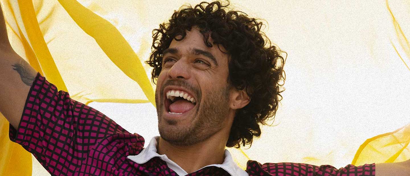 Foto mostra um homem branco, de cabelos ondulados curtos e de barba. Ele veste uma camiseta listrada vinho, grita e segura uma bandeira amarela.