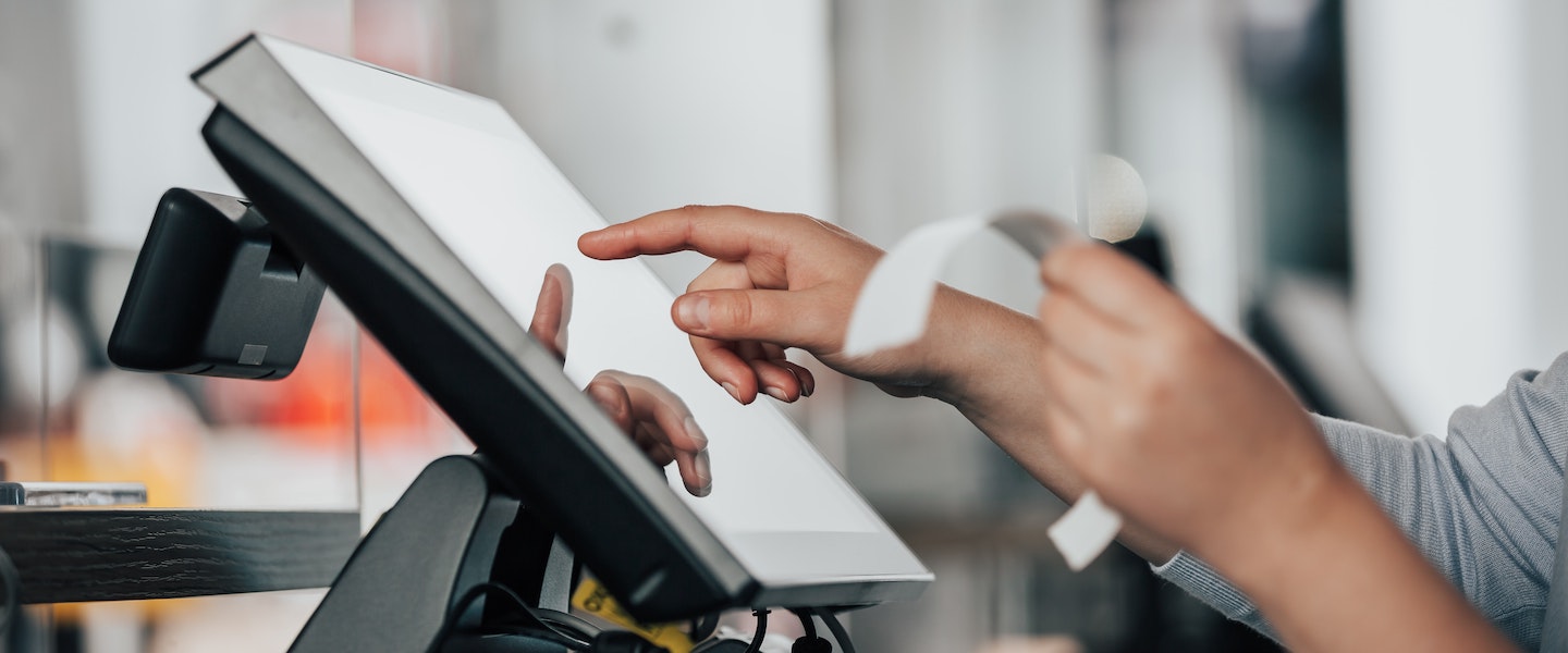 Imagem de uma mão segurando um recibo de compra e digitando num painel de caixa de loja.