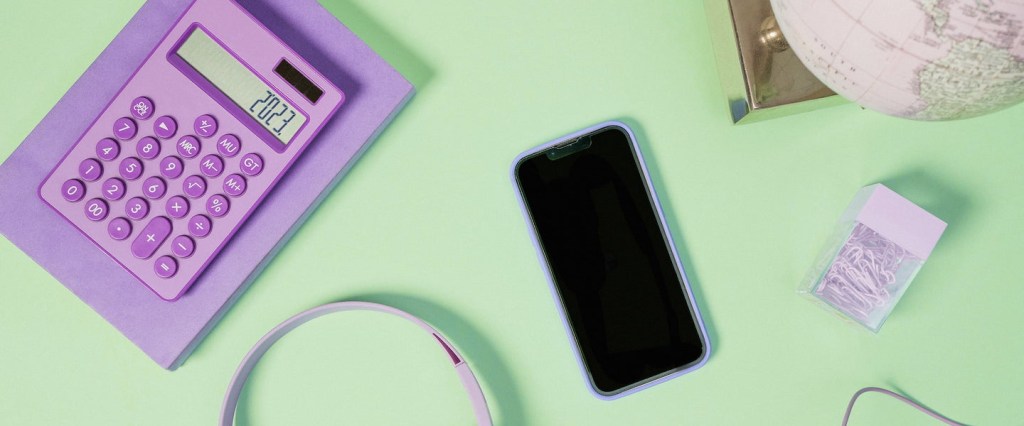 Imagem de uma calculadora lilás, com os números 2023 na tela, um celular desligado no meio, uma caixinha de clip, um globo terrestre e um headphone. Tudo numa superfície verde clara.