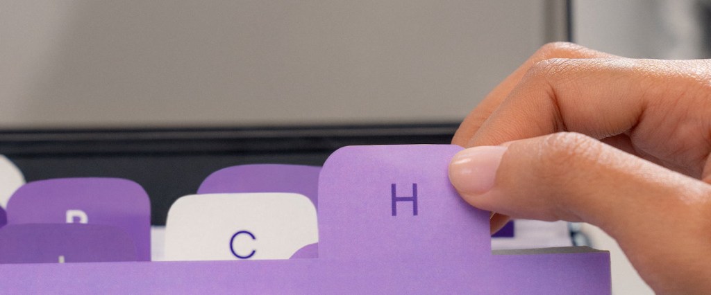 Imagem de uma mão segurando uma etiqueta roxa com a letra H dentro de um gaveteiro de escritório.