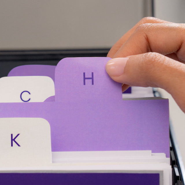 Imagem de uma mão segurando uma etiqueta roxa com a letra H dentro de um gaveteiro de escritório.