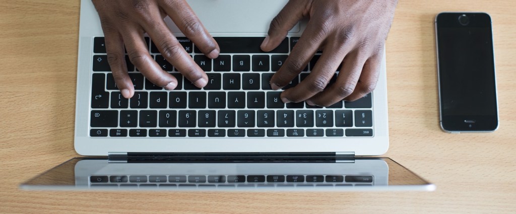 Imagem das mãos de um homem negro digitando em um computador. Ele usa relógio e veste camisa branca.