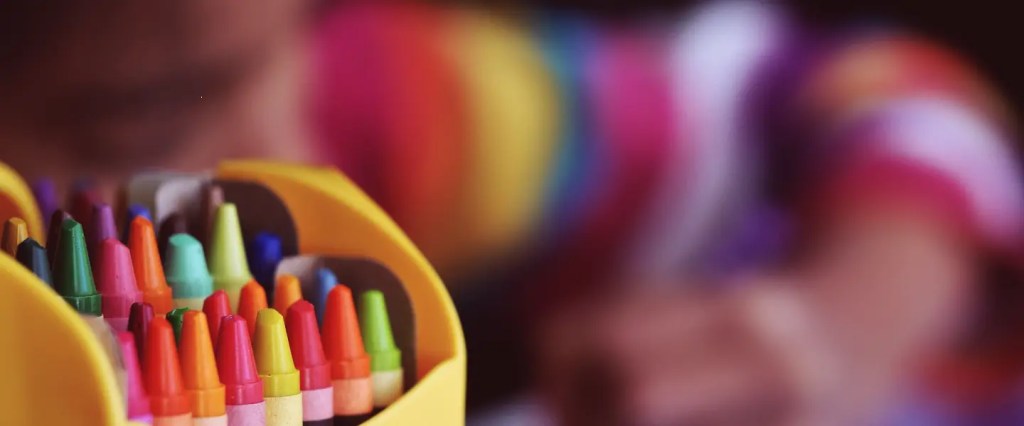 Criança desenhando ao fundo, com destaque para um caixa de giz de cera colorido ao lado esquerdo