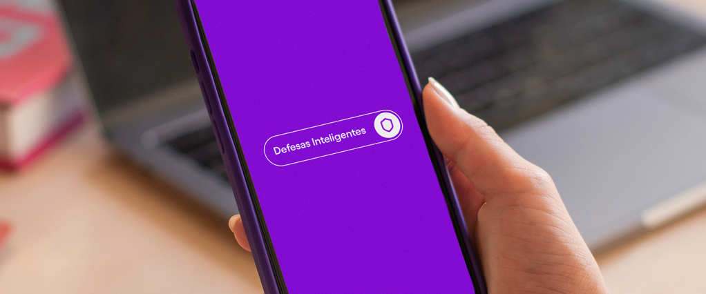 Imagem de uma mão segurando um celular. A tela é roxa e mostra a frase "Defesas Inteligentes" escrita em branco, ao centro.