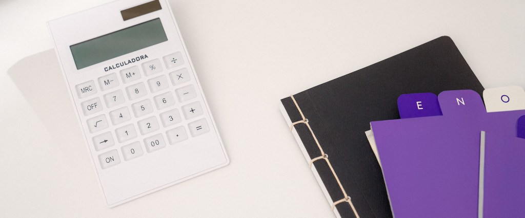 Foto de uma calculadora branca, ao lado de uma pasta preta e divisórias em tons de roxo e lilás, numa superfície clara.