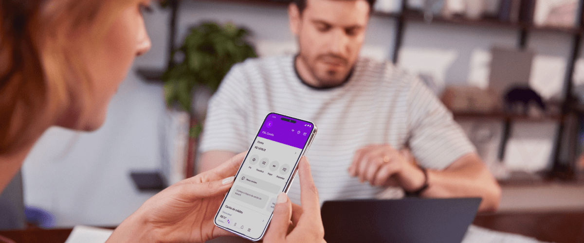 Informe de Rendimentos do Nubank: na imagem, uma mulher branca de cabelos loiros segura um celular, com a conta do Nubank aberta na tela, usando as duas mãos. Ao fundo, um homem branco está sentado à mesa olhando para um notebook preto.