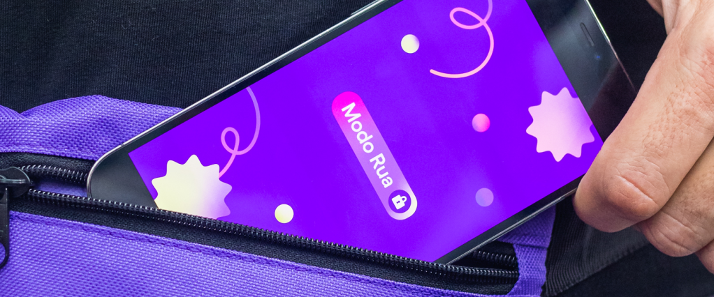 Imagem de um celular sendo retirado de uma pochete roxa. Na tela do celular, está escrito "Modo Rua" em um fundo roxo, com confetes de carnaval ao redor.