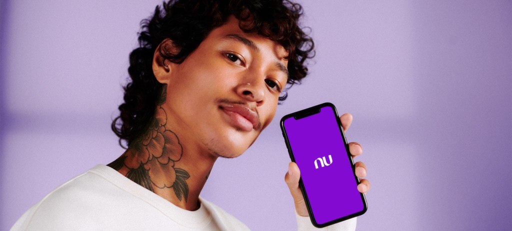 Um homem negro segurando um celular com o logo do Nu aparecendo. O fundo é lilás.