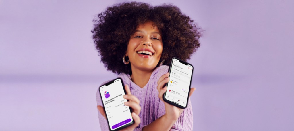 Imagem: ao centro do foto, uma mulher negra de pele clara, cabelos crespos e brincos de argola, em um fundo lilás, segurando dois celulares - um em cada mão -, em que mostram na tela o pagamento de iFood com NuPay.