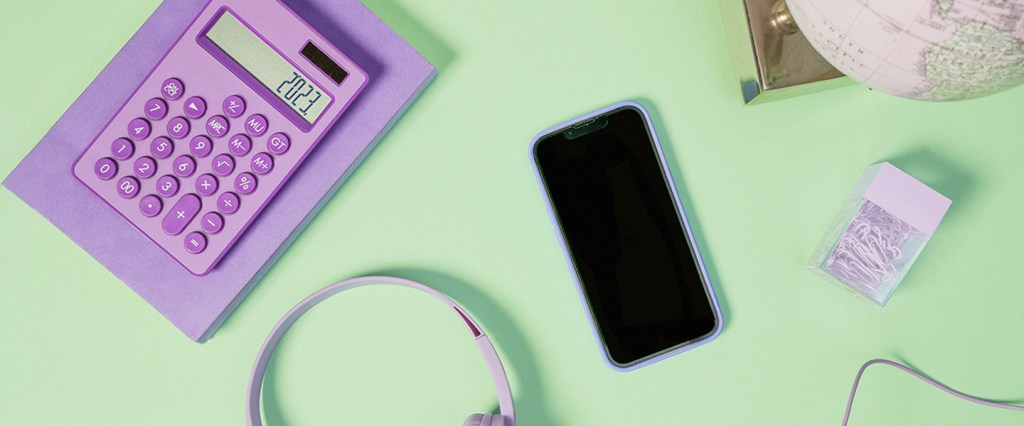 Imagem de um celular, uma calculadora roxa e um fone de ouvido roxo organizados aleatoriamente sobre um balcão verde. Há também uma caixa de clips brancos e um globo terrestre.