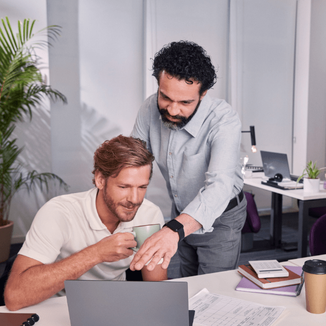 Dois homens brancos em um escritório olham para a tela de um notebook na mesa. Um deles tem cabelo preto e barba, está em pé, apontando para a tela. O outro está sentado, é loiro e segura uma xícara nas mãos.