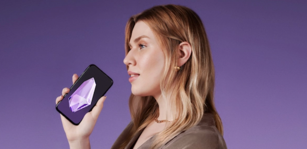 Foto de uma mulher loira virada de lado segurando um celular com a tela com um diamante (representando o pedacinho/ações do Nubank).