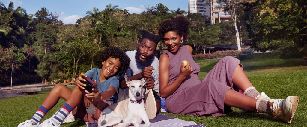 Imagem de uma família (um homem, um menino e uma mulher negros) sentados na grama de um parque. Tem um cachorro branco com eles.