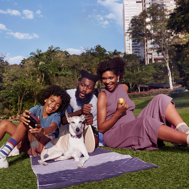 Imagem de uma família (um homem, um menino e uma mulher negros) sentados na grama de um parque. Tem um cachorro branco com eles.