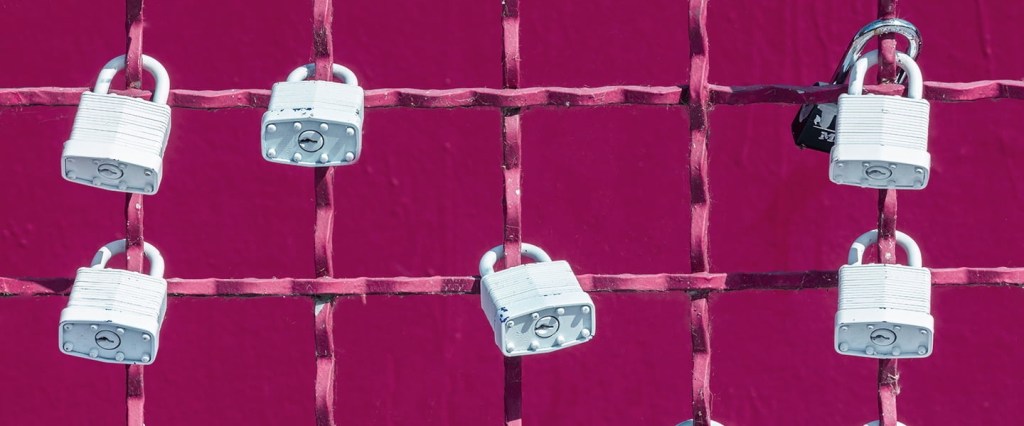 Imagem de cadeados brancos presos em uma grade na cor rosa magenta. O fundo da imagem também é rosa magenta.