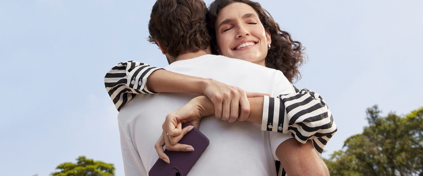 Imagem de uma mulher segurando um celular, sorrindo e abraçando um homem. Ele aparece de costas, usando camiseta branca.