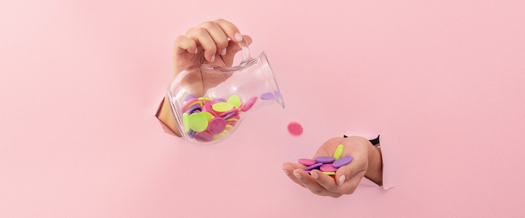 Foto de mãos ultrapassando um parede rosa clara, em que uma delas que segura moedas coloridas e, a outra, uma jarra transparente que despeja os objetos.